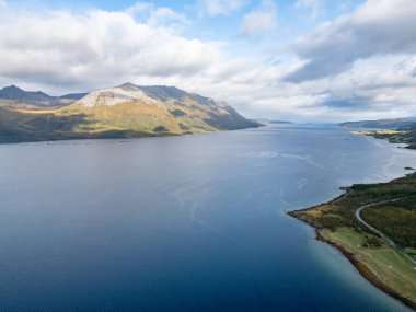 Kendinizi bu geniş hava fotoğrafıyla engin bir kuzey fiyordunun nefes kesici görkemine daldırın. Derin, sakin mavi sular ufka doğru uzanıyor, engebeli kaya yüzeylerini gösteren yüksek dağlarla çevrili..