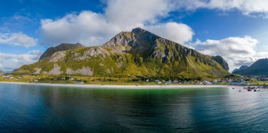 Norveç 'in panoramik fiyort manzarasının nefes kesici ihtişamını tecrübe edin. Bu geniş resim, el değmemiş beyaz kumsal ve sakin, derin mavi-yeşil sulara bakan görkemli, yüksek bir dağı gözler önüne seriyor..