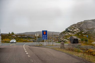 Norveç 'e uzak bir sınır geçişinin belirgin bir görüntüsü. Üzerinde Norveç arması olan belirgin bir mavi işaret var. Asfalt yol engebeli, kayalarla kaplı bir arazide kıvrımlar. Sonbaharda sarının gölgeleriyle süslenir..