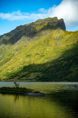 Daldır kendini canlı bir dağ gölünün dingin güzelliğine görkemli bir tepenin altında yemyeşil yamaçlarla. Güneş ışığı suyun yüzeyinde dans eder ve dokulu araziyi aydınlatarak büyüleyici bir ışık ve gölge oyunu yaratır.