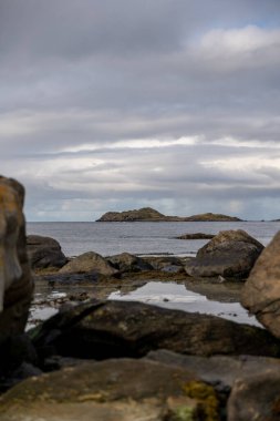Huzurlu ve atmosferik bir fotoğraf sakin bir kuzey kıyısı sahnesini yakalıyor. Ön plandaki büyük, yıpranmış kayalar bulutlu bir gökyüzünün sessiz ışığını yansıtan sakin bir su kütlesi. Orta mesafede, küçük, engebeli bir ada..