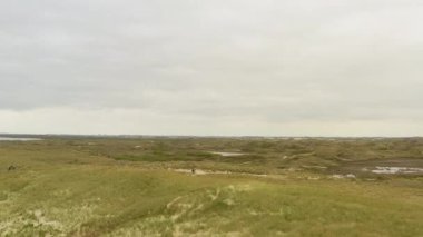 İHA 'lar büyük bir kumul üzerinde çekim yaparak bulutlu bir gökyüzünün altında, uzak bir yolda bisiklete binen iki kişiyi yakalıyorlar.