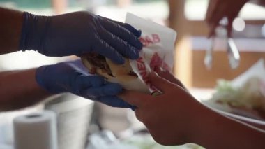 Bir şefin eldivenli ellerinin, hazır ve cömertçe hazırlanmış bir Dner Kebap sandviçini bir yemek standındaki müşteriye verirken çekilmiş yakın plan fotoğrafı.