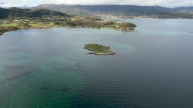 Sakin bir Norveç fiyordunun turkuaz sularında engebeli bir kıyı şeridi bulunan küçük, yemyeşil bir adanın detaylı hava görüntüleri..