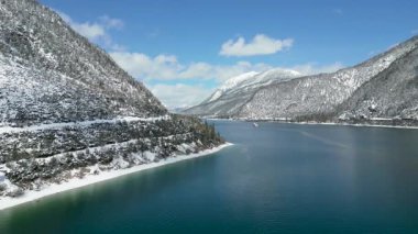 Muhteşem bir turkuaz dağ gölü üzerinde seyreden yolcu feribotunun hava görüntüsü, görkemli karla kaplı tepeler ve canlı mavi bir gökyüzü..