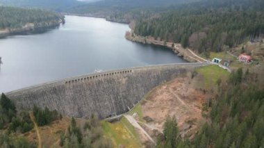 Geniş ve yavaş bir drone, bulutlu bir gökyüzünün altında sakin bir gölü olan ormanlık bir vadide büyük eğimli bir taş barajın görüntüsü..