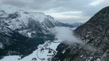 Karla kaplı bir köyü ve yükselen tepeler arasında kıvrılan bir yolu olan görkemli bir dağ vadisinde nefes kesici bir hava manzarası..