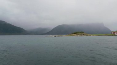 İskandinavya 'da sakin bir fiyort manzarası, küçük, kayalık bir ada ve sağda geleneksel kırmızı bir ev, dramatik, bulutlu bir gökyüzünün altında..