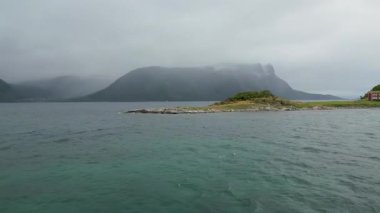 İskandinavya 'da sakin bir fiyort manzarası. Küçük, kayalık bir ada ve bulutlu bir gökyüzünün altında geleneksel kırmızı bir ev..