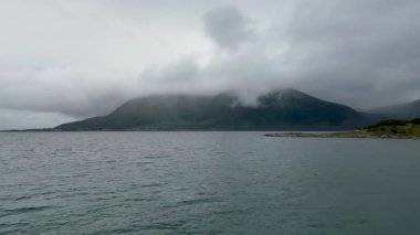 İskandinavya 'da sakin bir fiyordun manzarası. Küçük, kayalık bir ada ve bulutlu bir gökyüzünün altında büyük, puslu bir dağ..