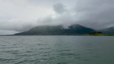 İskandinavya 'da sakin bir fiyordun manzarası. Küçük, kayalık bir ada ve bulutlu bir gökyüzünün altında büyük, puslu bir dağ..