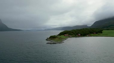 Engebeli bir kıyı şeridi, geleneksel kırmızı evler kümesi ve uzak mesafedeki bir teknenin yer aldığı nefes kesici İskandinav fiyortlarının insansız hava aracı görüntüleri..