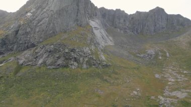 Majestic Lofoten Adaları Manzarası: İnsansız hava aracı görüntüsü, yumuşak, bulutlu bir gökyüzü altında keskin, kayalık zirvesi ve canlı yeşil yamaçlarıyla Norveç dağının engebeli güzelliğini gözler önüne seriyor.