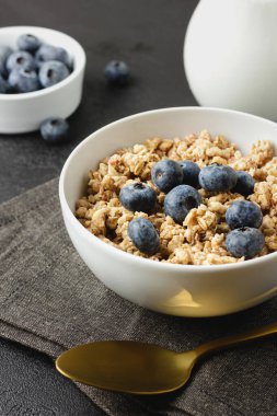 Granola with blueberry in white bowl on dark napkin. Healthy food and milk on black table with golden spoon. Side view of homemade breakfast.