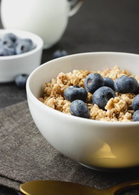 Granola with blueberry in bowl on dark napkin. Healthy breakfast on black table with golden spoon. Side view of homemade food.