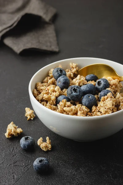 Granola with blueberry in white bowl. Healthy breakfast on black table with cotton napkin. Side view of homemade food.