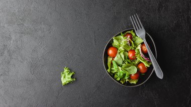 Green salad leaves with cherry tomatoes in bowl on black table. Fresh and healthy food with copy space. Top view.