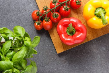 Ağaç kesme tahtasında biberli ve fesleğenli kiraz domatesleri. Gri masada taze sebzeler ve fotokopi alanı. Üst görünüm.