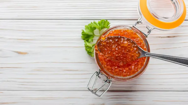 Red caviar with parsley in jar and spoon. Seafood with copy space on white wooden table. Luxury salmon delicacy. Top view.