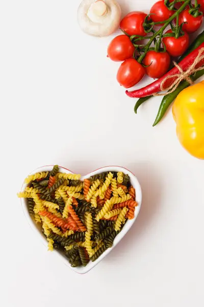 Çeri domatesli, kırmızı biberli, mantarlı ve biberli kalp kasesinde çiğ makarna. Çoklu renkli tortinin üst görünümü.