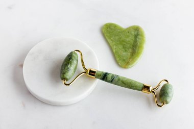 Green jade roller and gua sha stone massager for face on white marble. Trendy cosmetic tool. Anti aging therapy. Side view. Flat lay.  clipart