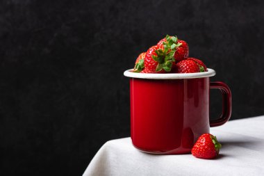 Emaye bardakta taze çilek. Beyaz masa örtüsünde kırmızı böğürtlen ve siyah duvar. Sağlıklı ve organik tatlı..