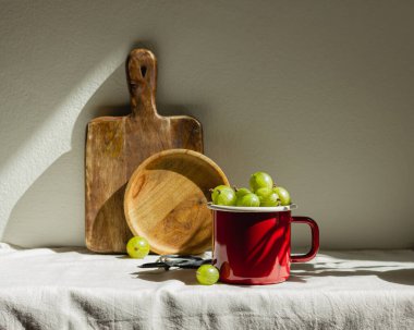 Bektaşi üzümü, kırmızı bir diş minesi bardağında, gün ışığında. Açık masa örtüsü üzerinde taze yeşil meyveler, ahşap tahta ve tabakla..
