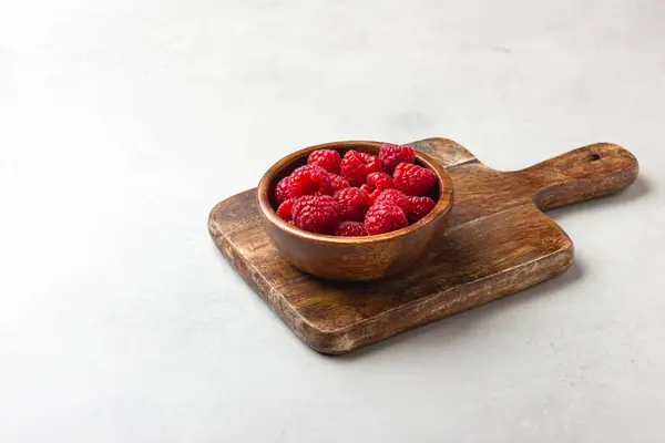 Ahşap bir kasede taze ahududu ve açık gri arka planda bir kesim tahtası. Kırmızı çilek.