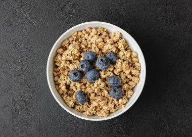 Beyaz kasede yaban mersinli granola. Siyah masada sağlıklı bir kahvaltı. Ev yapımı yiyeceklerin en üst görüntüsü.