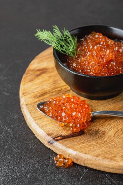Kavanozda dereotu olan kırmızı somon havyarı. Siyah arka planda gümüş kaşıklı deniz ürünleri. Tahta kesme tahtasındaki lüks lezzet. Yan görünüm.
