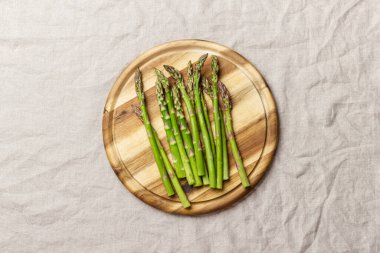 Yuvarlak ahşap bir tahta üzerinde yeşil kuşkonmaz keten bir masa örtüsünün üzerinde. Taze ve doğal pişmemiş kuşkonmaz. Sağlıklı bitki yiyeceği.