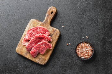 Raw beef meat on a wooden cutting board. Three pieces of fresh uncooked red meat on dark background with pink salt. Top view.