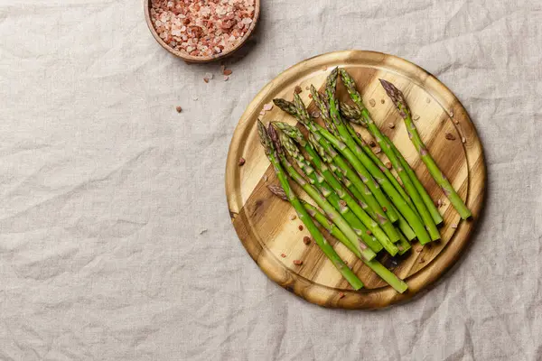 Ahşap bir masa örtüsünün üzerinde pembe tuz ve yeşil kuşkonmaz. Taze ve doğal pişmemiş kuşkonmaz. Sağlıklı bitki yiyeceği.