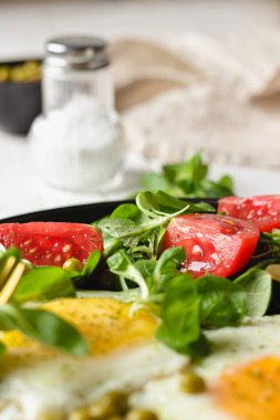 Tavada vişneli domatesli yumurta ve salata. Küçük kasede yeşil bezelyeler, arka planda tuz ve bej peçete. Beyaz ahşap masada ev yapımı kahvaltı. Yan görünümü kapat.