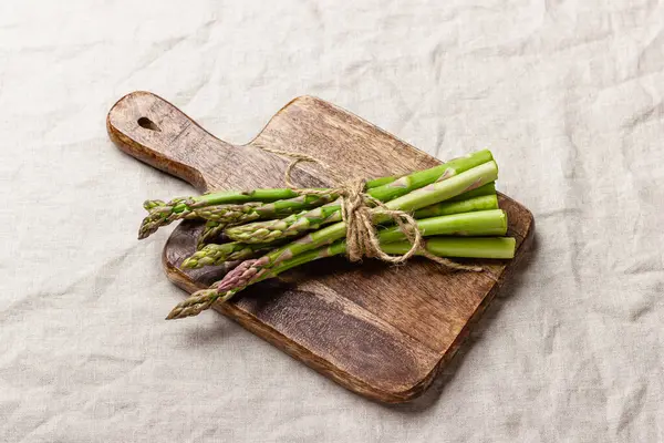 Keten masa örtüsünün üzerindeki tahta kesme tahtasındaki yeşil kuşkonmaz. Taze ve doğal pişmemiş kuşkonmaz. Sağlıklı bitki yiyeceği.
