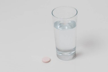 İçme suyuyla dolu şeffaf bir bardak. Su bardağının yanında yuvarlak bir vitamin tozu var. Arkaplan beyaz.