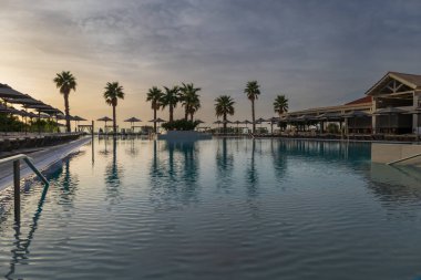 Havuzun üzerinde gün doğumu ve güneş yatakları. Deniz kıyısının arka planında. Büyük palmiye ağaçları.