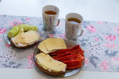 Kahvaltı mutfak masasının üstünde. Salamlı ve peynirli ekmek. Garnitür sebzedir, salatalık ve biber. Bardakta çay.