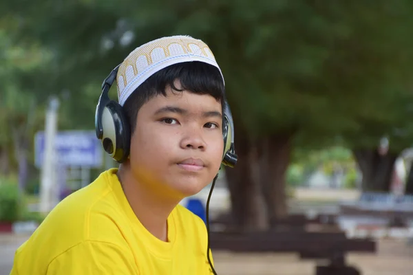 ポートレート黄色のシャツと帽子を身に着けている若いアジアのイスラム教徒の少年は ヘッドフォンを保持し 公園の木の下に座って 柔らかくて選択的な焦点 — ストック写真