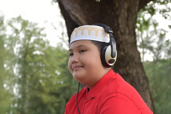 年轻的亚洲穆斯林男孩穿着红衫 头戴帽子 头戴帽子 手持耳机 坐在公园的树下 目光柔和而有选择性 — 图库照片