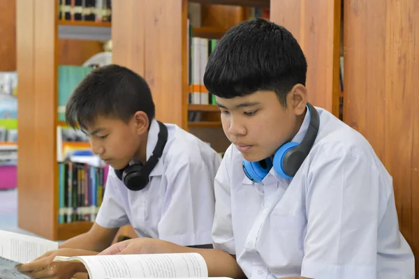 Soft fucus of two asian boy students are listening media, reading and consulting about favourite book at library of school
