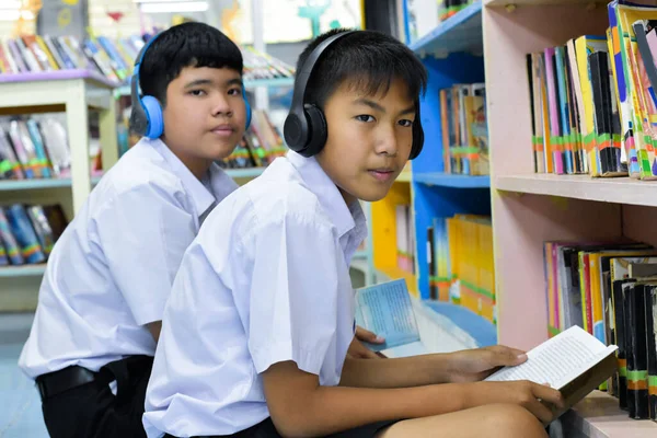 Soft fucus of two asian boy students are listening media, reading and consulting about favourite book at library of school