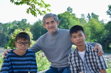 An elderly asian male is spending his spare time explaining the contents of the lesson and happily helping his two grandsons with their homework and school projects in his backyard, soft focus.