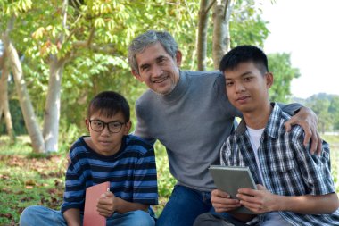 An elderly asian male is spending his spare time explaining the contents of the lesson and happily helping his two grandsons with their homework and school projects in his backyard, soft focus.