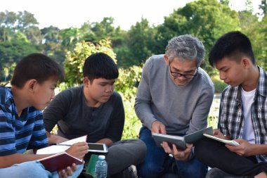 An elderly asian male is spending his spare time explaining the contents of the lesson and happily helping his two grandsons with their homework and school projects in his backyard, soft focus.