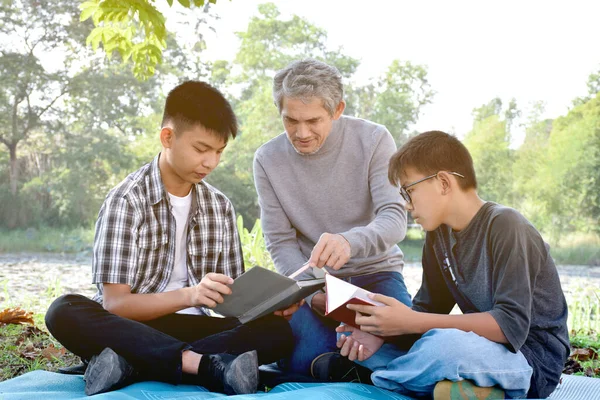 Asian elder and grandsons are spending time together in the park by teaching, reading and talking about topic in the book which they are interested in happily, soft  and selective focus on first boy.