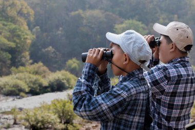Asian boys are using binoculars to do the birdwatching in tropical forest during summer camp, idea for learning creatures and wildlife animals and insects outside the classroom.