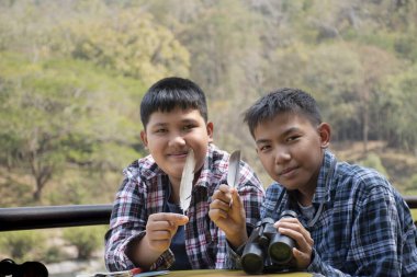 Asian boys hold bird feather and binoculars sit together in local national park to learn differentces of birds from feather during their summer vacation, leisure and outdoor activity of teens concept.