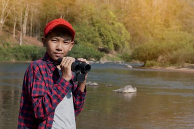 Young asian boy in plaid shirt and red cap holds binoculars in hands and watching the fish in the river during his vacation and his school project work in summer, soft focus, nature learning concept.