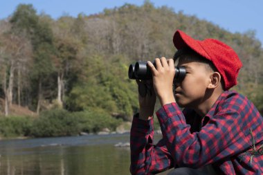 Young asian boy in plaid shirt and red cap holds binoculars in hands and watching the fish in the river during his vacation and his school project work in summer, soft focus, nature learning concept.